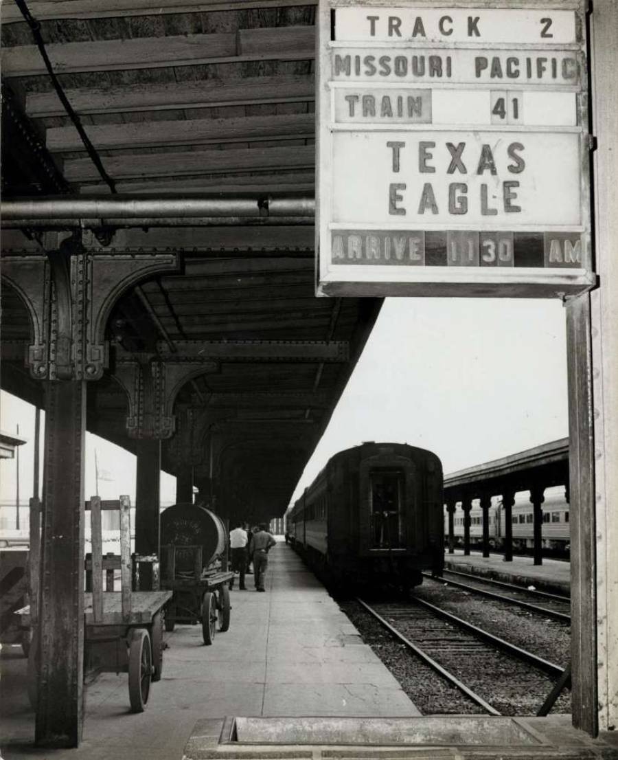 Black and white photo of a train station platform with a sign reading "Missouri Pacific Train 41 Texas Eagle Arrive 11:30 AM." A train and a few people are visible on the platform.