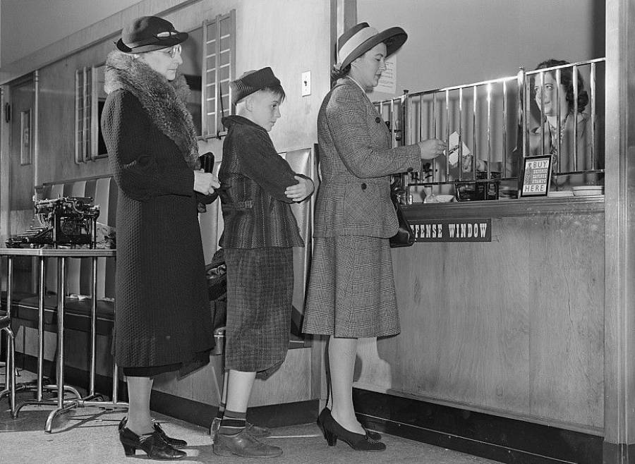 Three people in vintage clothing stand in line at a ticket window, while a woman behind the counter assists them. There is a typewriter on a nearby table and a "Pay Here" sign visible.