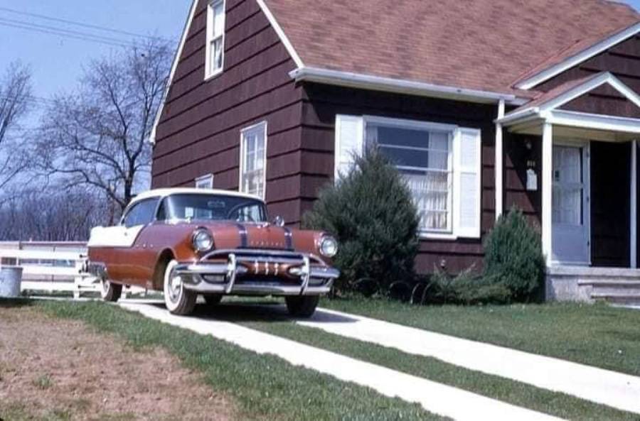 A vintage car is parked on the driveway of a brown house with white trim, green shrubs, and a sloped roof, on a sunny day with clear skies and bare trees in the background.
