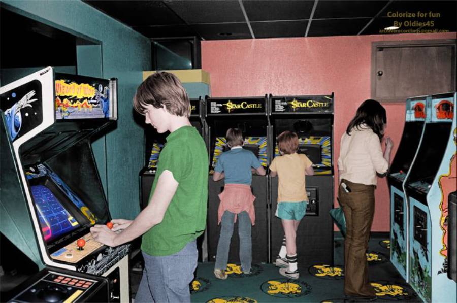 Four kids play at vintage arcade machines in a retro game room. Three face the machines, while one boy in a green shirt plays a game near the camera. The room has bright colors and classic arcade cabinet designs.