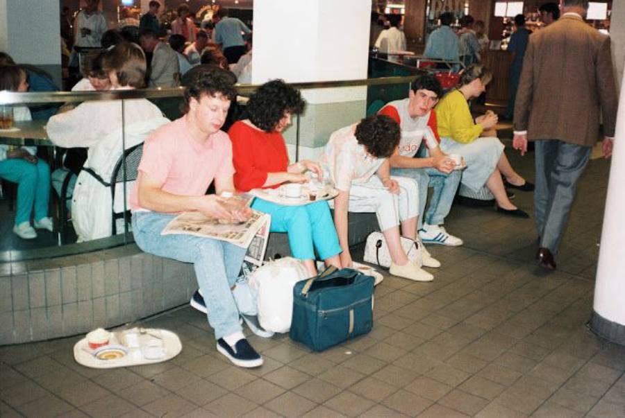 A group of people sit on benches in a busy indoor public area, eating and reading. Some have food trays and bags near them. Other people are walking or sitting in the background. The scene appears casual and crowded.