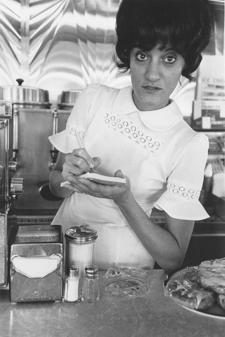 A waitress in a white uniform stands at a diner counter, holding a notepad and pen, looking up while taking an order. Metal appliances and condiments are visible on the counter.