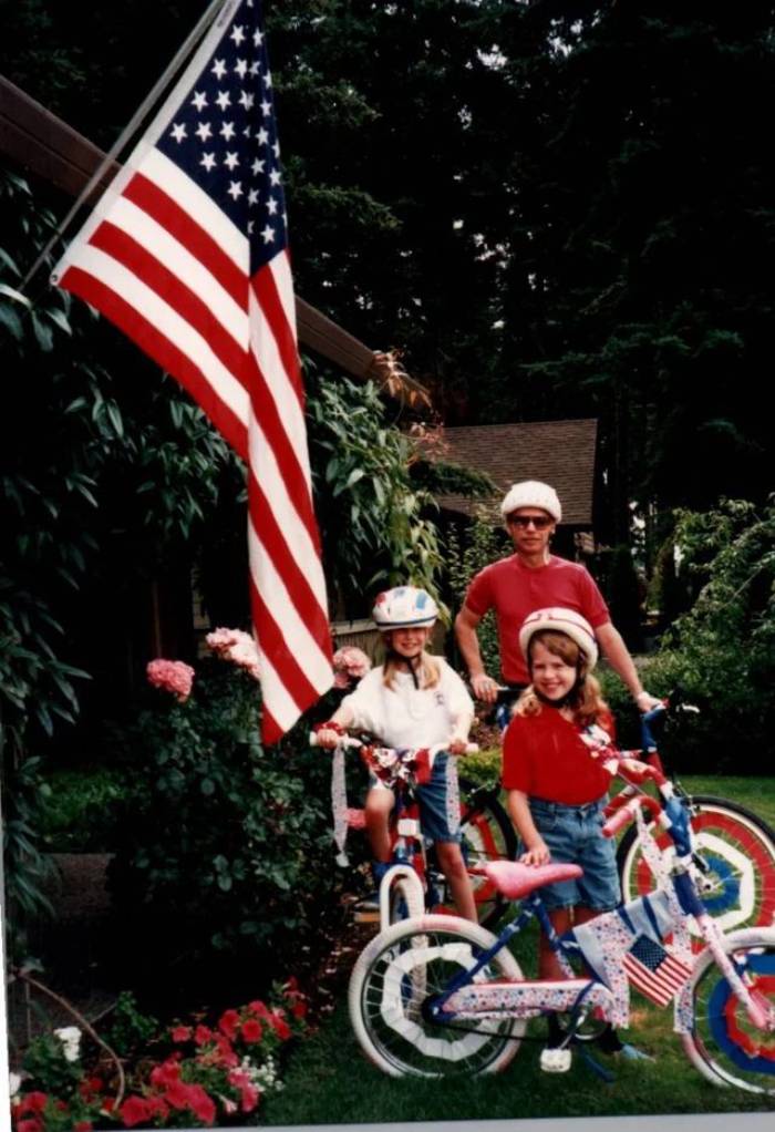 Three people, two children on decorated bikes and an adult standing behind them, pose outdoors near a large American flag. All wear helmets and casual clothes, surrounded by greenery and flowers.