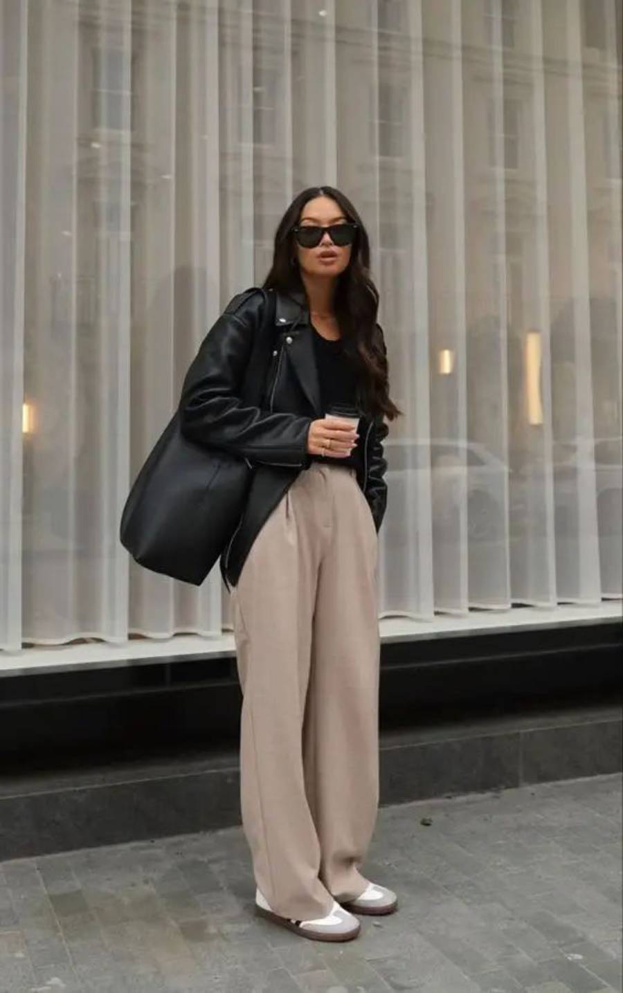 A woman wearing a black leather jacket, black top, tan wide-leg pants, and white sneakers stands holding a coffee cup and a large black bag. She has long hair, wears sunglasses, and stands in front of a sheer-curtained window.