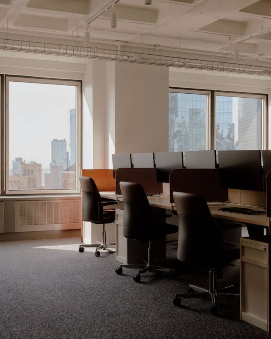 A modern office space with several empty desks and black chairs near large windows, overlooking city buildings on a bright day.