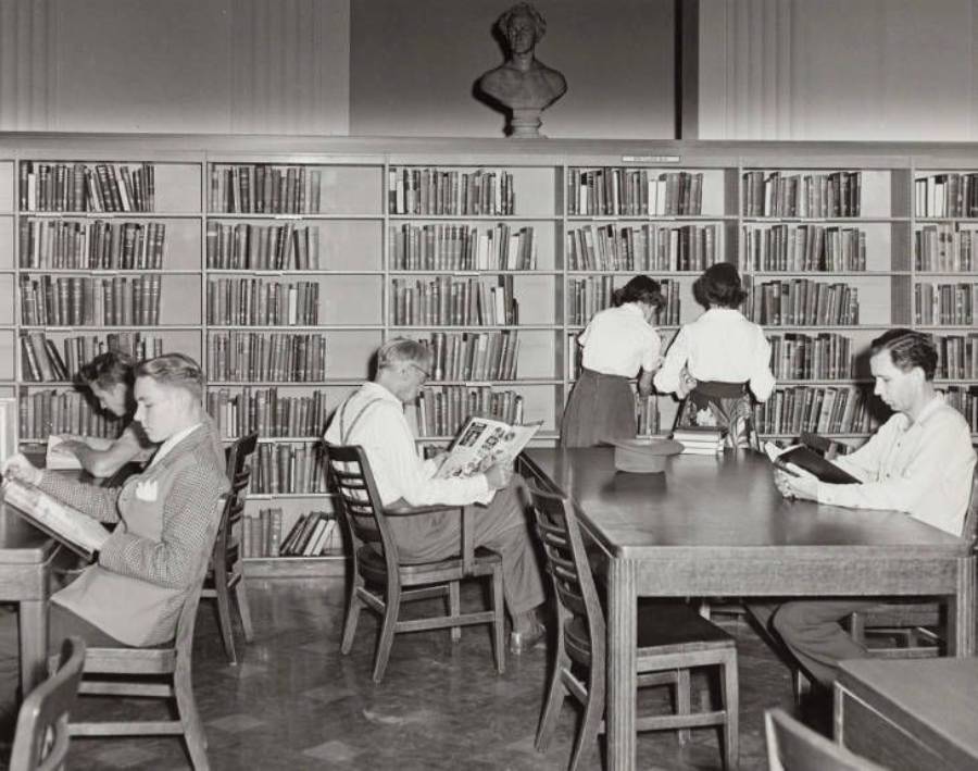 Black and white photo of people reading or browsing books in a library. Four people sit at tables reading, while two stand near tall bookshelves. A sculpture bust is on top of the shelves in the background.