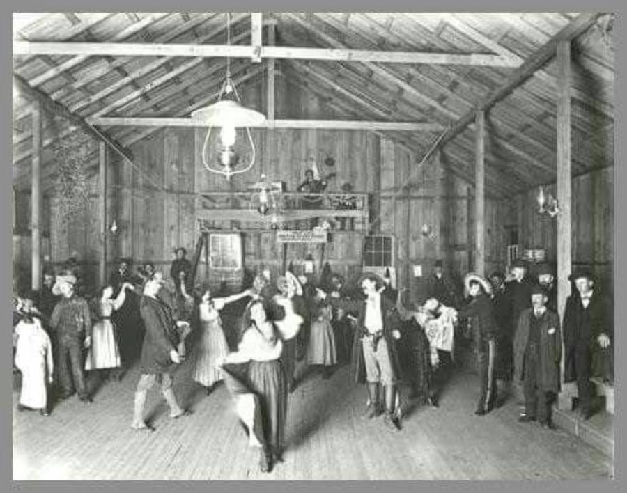 Black and white photo of people dancing inside a wooden hall, with musicians on a raised platform at the back. Men wear suits and hats; women wear dresses. The hall has exposed beams and hanging lights.