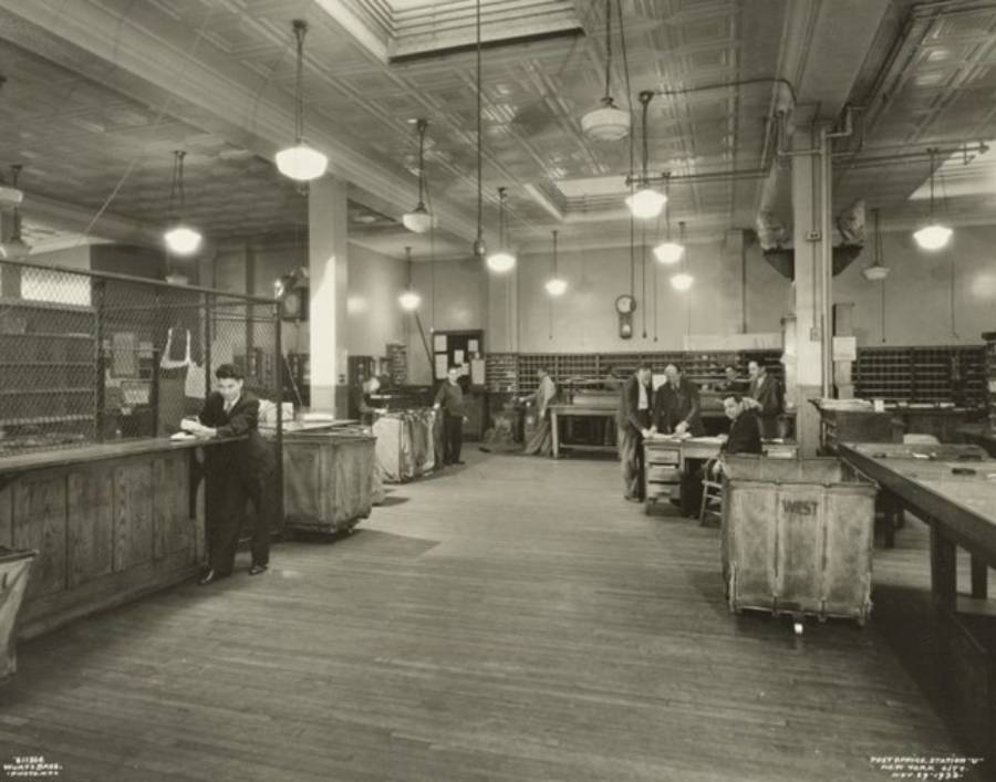 Black-and-white photo of a spacious mailroom with high ceilings. Several workers sort and process mail at tables and sorting bins. Overhead lights illuminate the room.