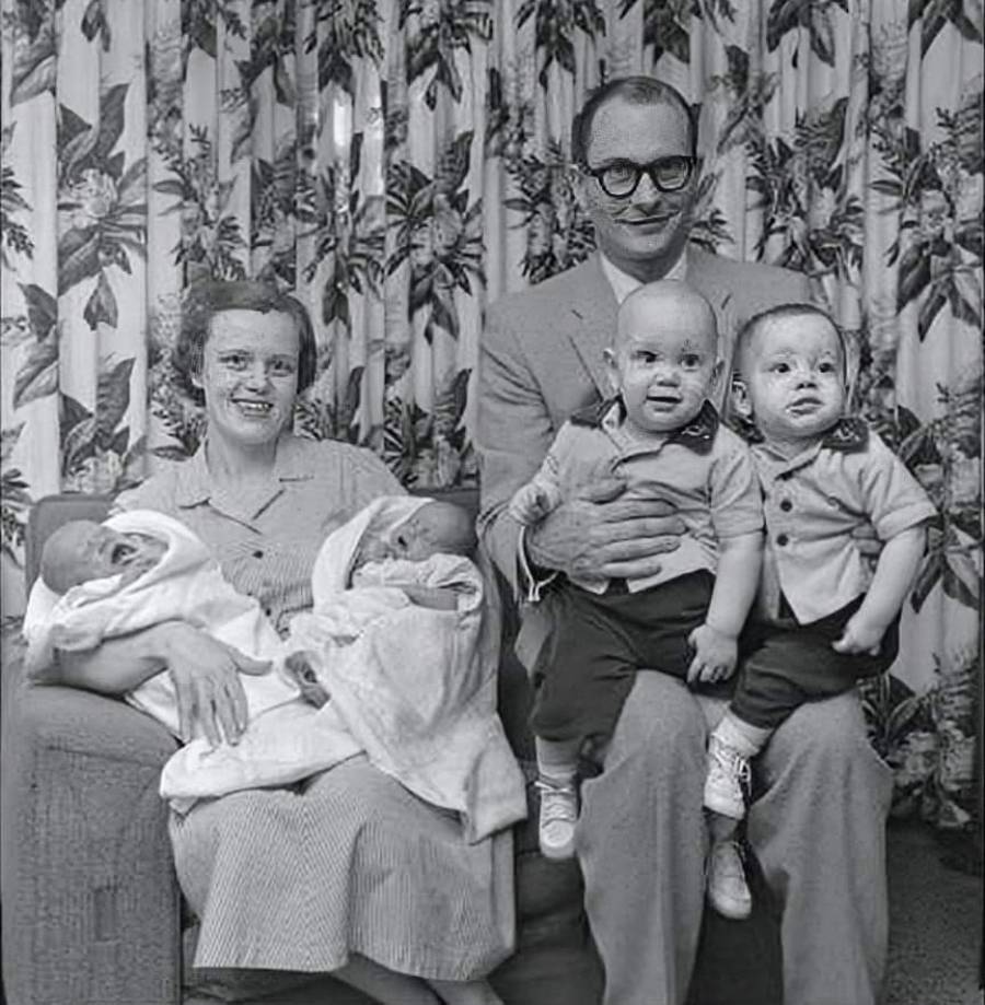 A black-and-white photo of a smiling couple sitting with four young children: the mother holds two swaddled babies, while the father holds two toddlers on his lap, all posed in front of a floral curtain.