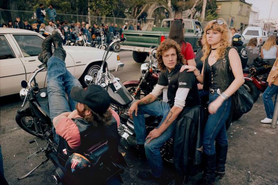 Several people in biker attire sit and stand around motorcycles on a busy street, with a crowd and parked cars in the background. One man reclines on his bike, while a woman stands beside another seated man.