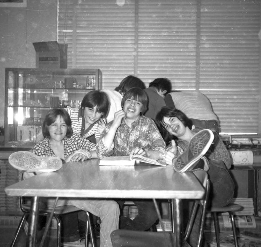Five kids sit and laugh around a classroom table, some with feet up, while two others face away at the back. A cabinet and large window with blinds are in the background. The mood is playful and relaxed.