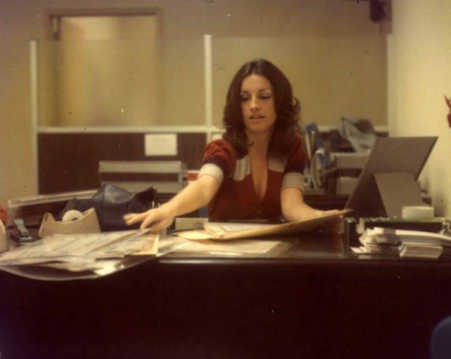 A woman with long brown hair sits at a cluttered office desk, reaching out to organize papers. She is wearing a red top with light-colored accents. There are folders, documents, and office equipment around her.