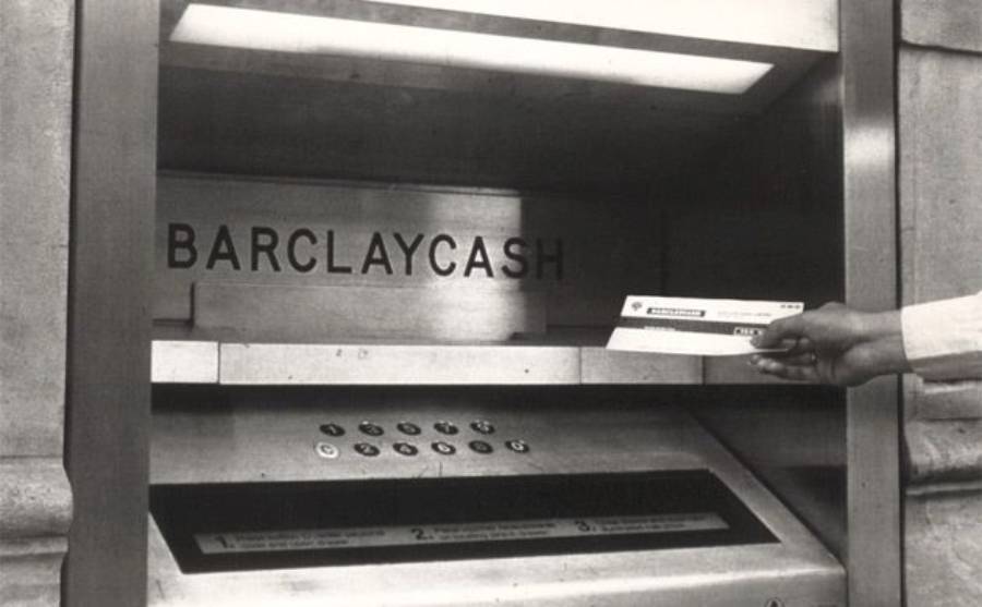 A person inserts a paper voucher into a vintage BARCLAYCASH ATM machine, with buttons and instructions visible below the slot.