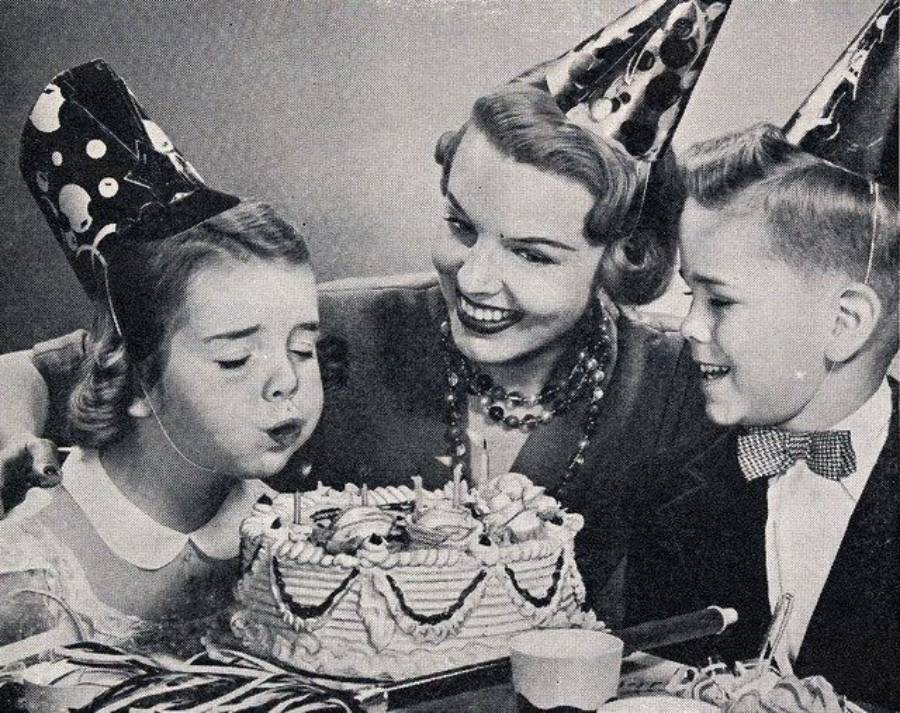 A girl blows out candles on a birthday cake, sitting between a smiling woman and a boy. All three wear party hats and appear happy. The photo is in black and white.