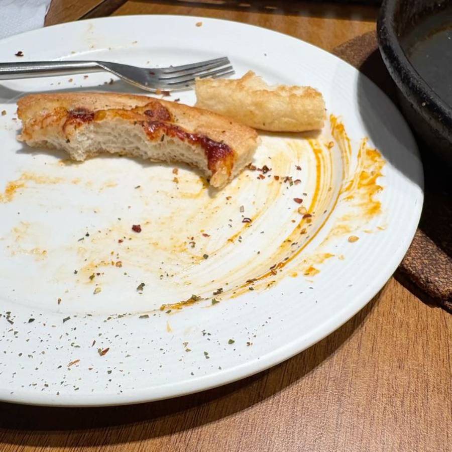 A mostly empty white plate with pizza crust, a breadstick, a fork, and sauce stains. Part of a dark pan is visible to the right, all on a wooden table.