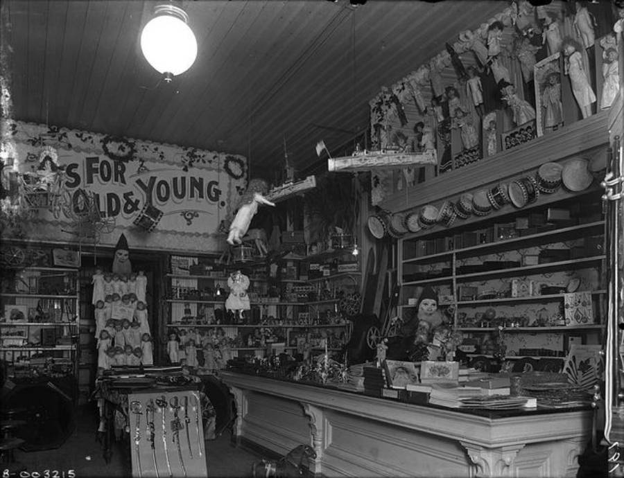 A vintage toy shop filled with dolls, toys, and shelves of various items. A sign reads “For Old & Young.” A person stands behind a counter, surrounded by merchandise, under a hanging light.