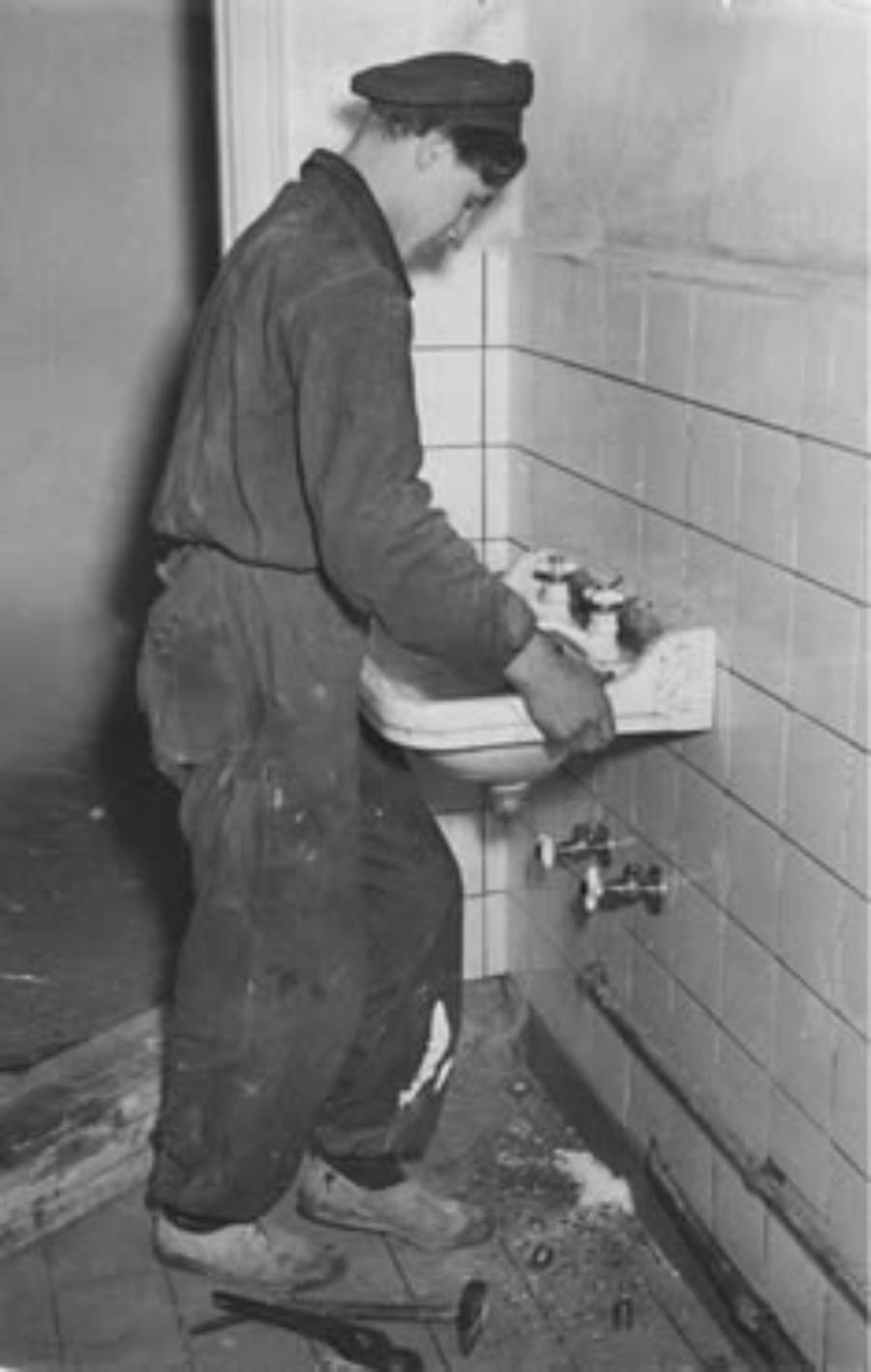 A person in work clothes and a cap is repairing or installing a wall-mounted sink in a tiled room, with tools and debris on the floor nearby.