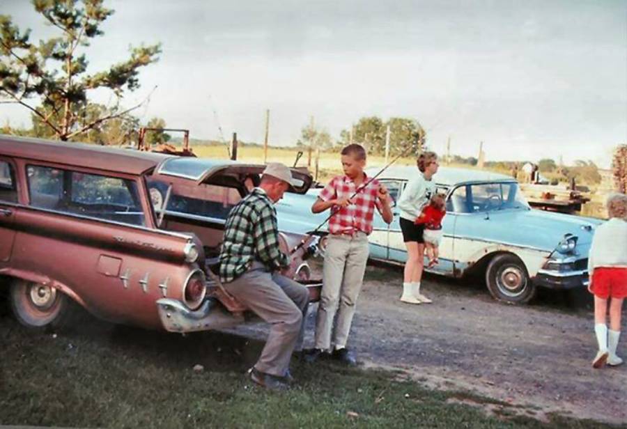 Two boys examine fishing poles near an old pink station wagon, while a woman and two children stand by a light blue vintage car in a rural outdoor setting.