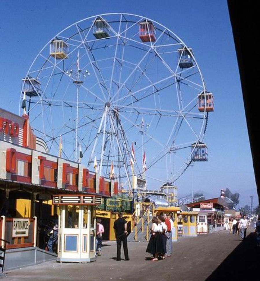 A vintage Ferris wheel with colorful gondolas stands at a fairground. People gather near ticket booths and food stalls under a clear blue sky. The scene has a nostalgic, mid-20th-century feel.