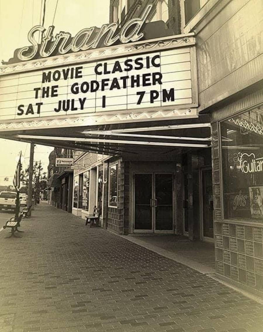 Sepia-toned photo of the Strand theater marquee announcing "Movie Classic: The Godfather, Sat July 1, 7PM" on a brick sidewalk with benches and storefronts visible.