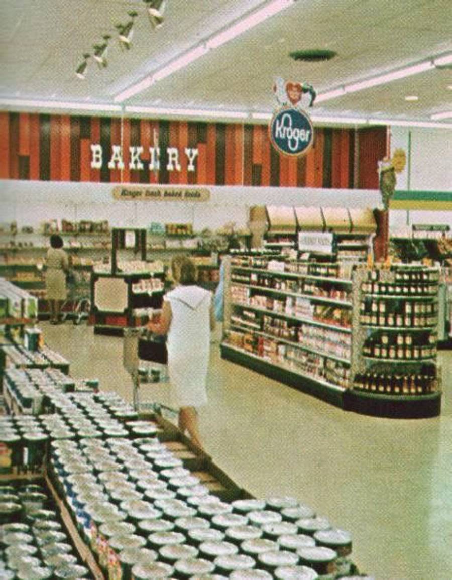A woman in a white dress shops in a vintage Kroger grocery store, heading toward the bakery section. Shelves filled with canned goods and various products line the brightly lit, retro-style store.