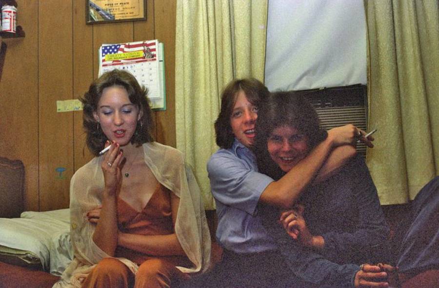 Three young people sit indoors; a woman in a dress smokes on the left while two others hug and smile on a bed. There’s wood-paneled walls, a calendar, curtains, and a window air conditioner in the background.