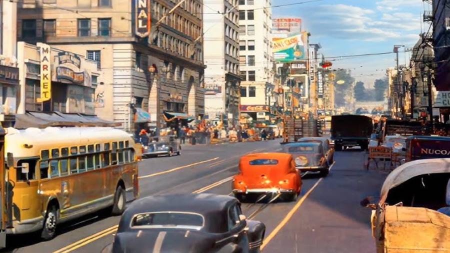 A busy city street scene from the mid-20th century with vintage cars, a yellow bus, streetcars, storefronts, pedestrians, and tall buildings under a blue sky.