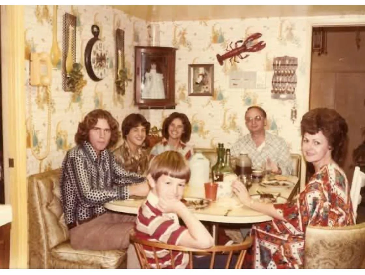 Six people sit around a dining table in a vintage kitchen with patterned wallpaper, decorative items, and a large fake lobster on the wall. Plates, drinks, and food are on the table as they smile at the camera.