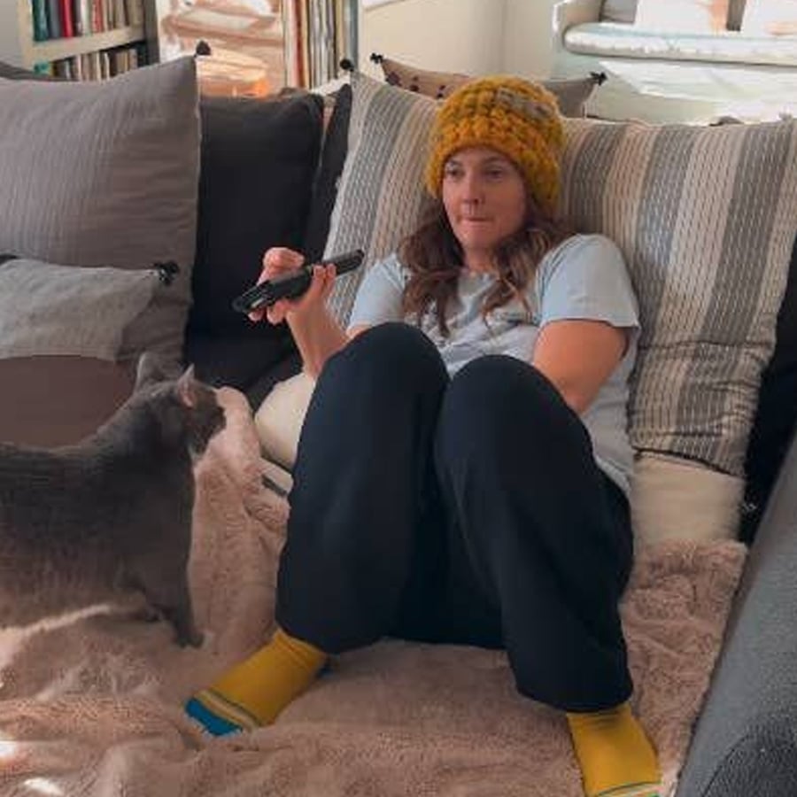 A person wearing a yellow knit hat and yellow socks sits on a couch holding a TV remote. A gray cat stands nearby on the couch, and a blanket covers part of the person’s legs.