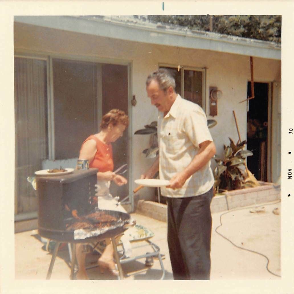 A man grills meat while a woman stands nearby in a sunlit backyard patio with plants and a sliding glass door. The photo has a vintage look.