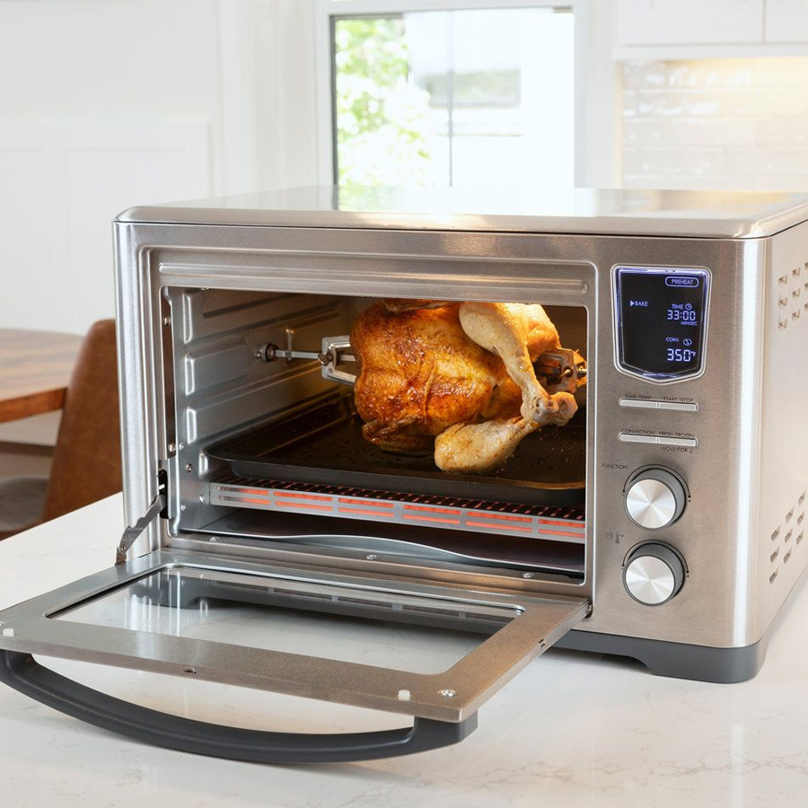 A stainless steel countertop oven with digital controls is cooking a whole rotisserie chicken. The oven door is open, and the chicken is turning on a spit inside.
