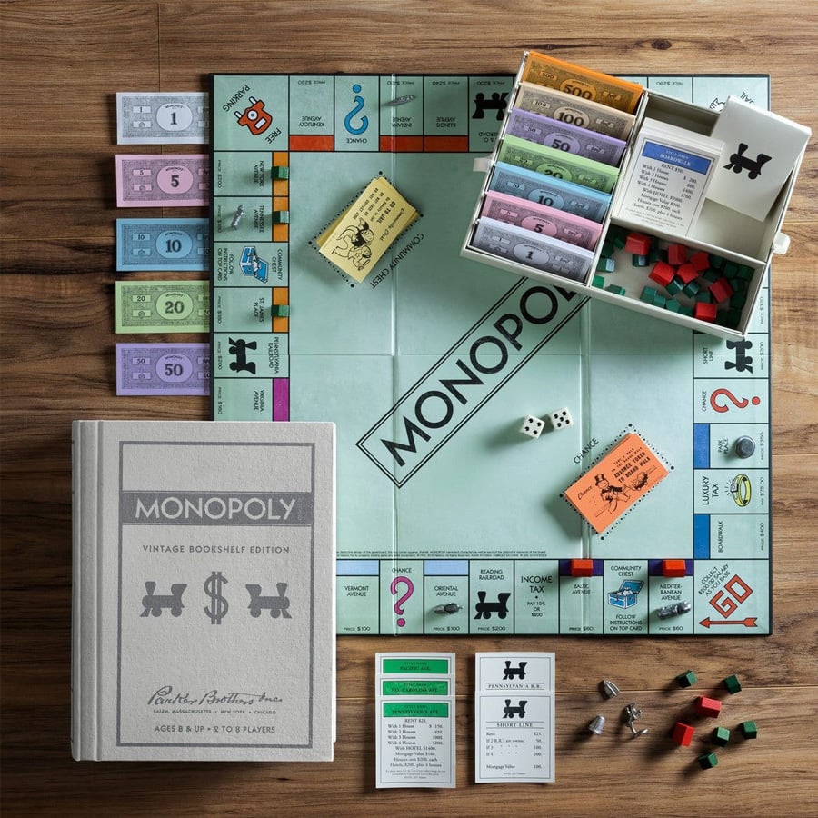 Monopoly board game setup with game pieces, cards, organized money, houses, and hotels. The vintage edition book-style box is closed and lies beside the open board on a wooden surface.