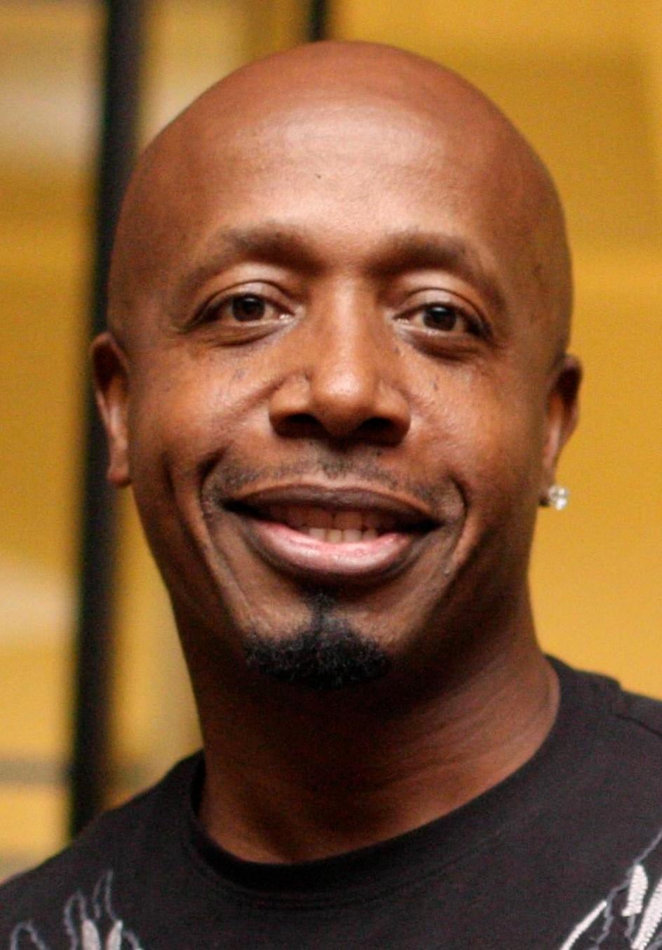 A smiling bald man with a trimmed goatee and earring, wearing a black shirt, stands in front of a blurred yellow background.