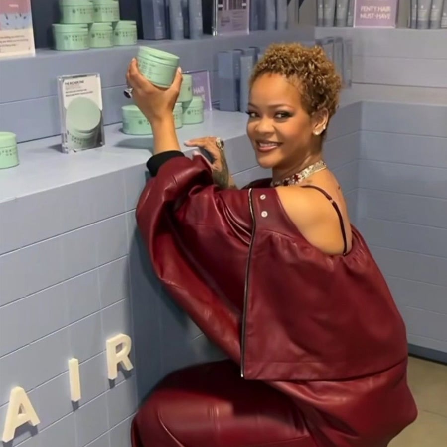 A woman in a maroon outfit smiles while crouching by a display of round mint-green product jars, holding one up in her hand in a modern store setting.
