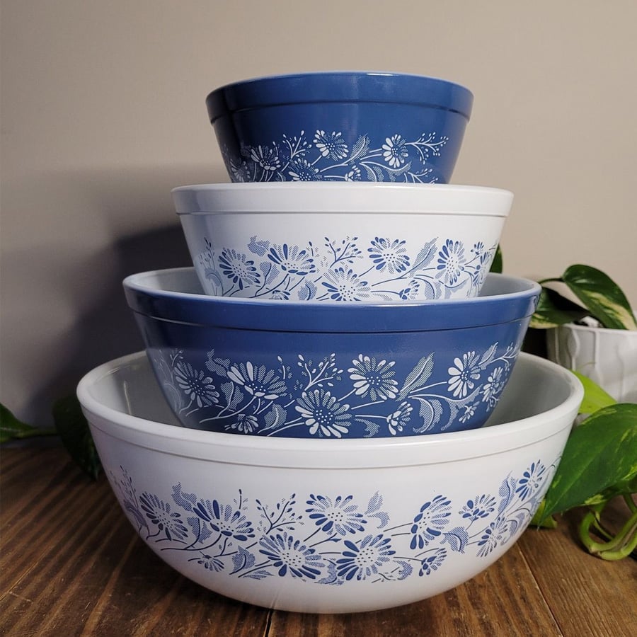 Four vintage mixing bowls, alternating blue and white, are stacked inside each other. Each bowl has a delicate white or blue floral pattern. The stack sits on a wooden surface with green leaves nearby.