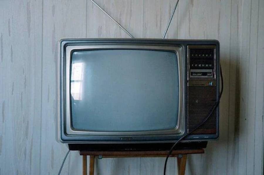 A vintage television with a curved screen, two antennae on top, and dials on the side sits on a small wooden stand against a pale, textured wall.