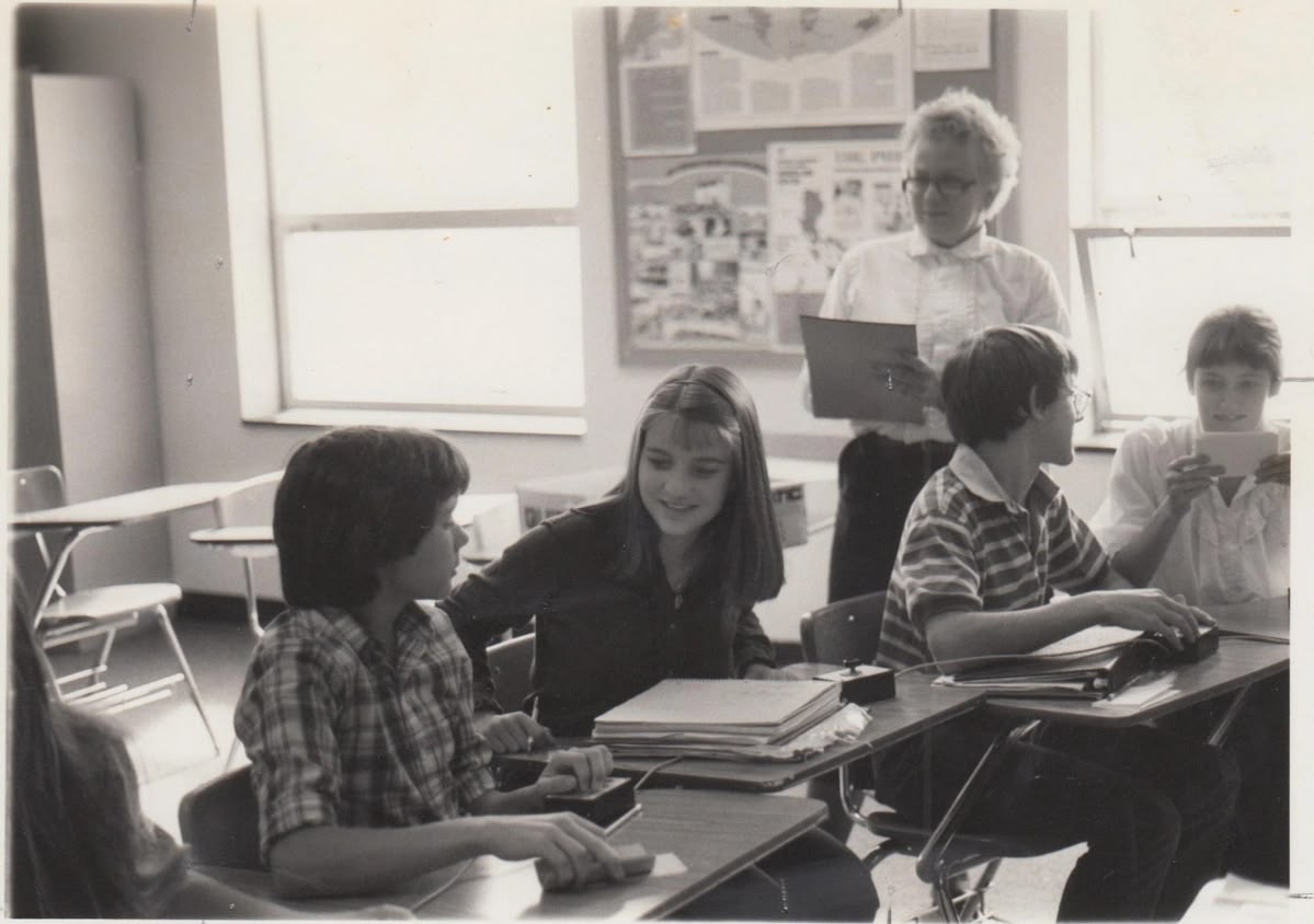 Black and white photo of students sitting at desks in a classroom, talking and smiling. An adult stands behind them, holding papers, appearing to interact with the students. Posters are visible on the walls.