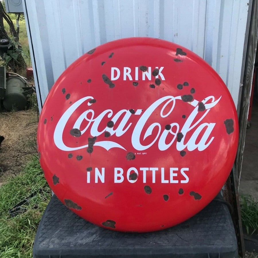 A round, red, weathered Coca-Cola sign reads "Drink Coca-Cola in Bottles" in white letters, leaning against a metal wall.
