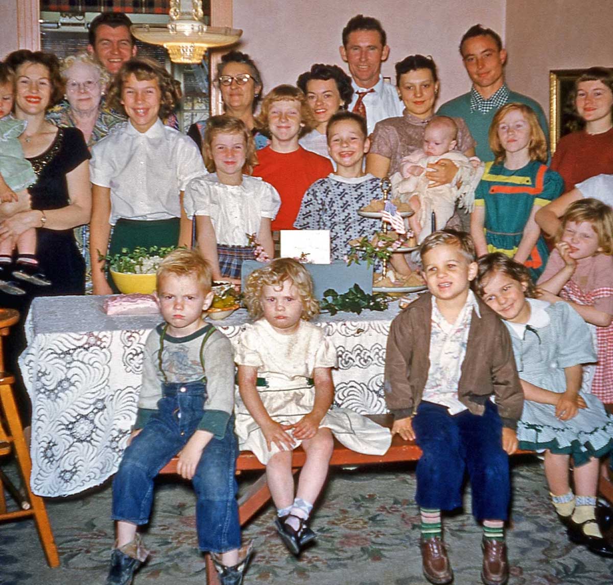 A large family group, including adults and children, poses indoors behind a table with a lace tablecloth and food. Everyone is dressed in vintage clothing, and some children are sitting in front.