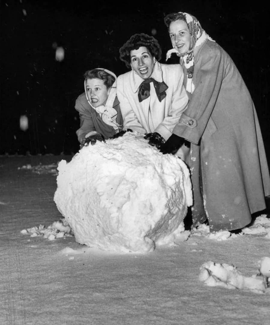 Three women, dressed in winter coats and scarves, smile and laugh while rolling a large snowball together at night in a snowy outdoor setting.
