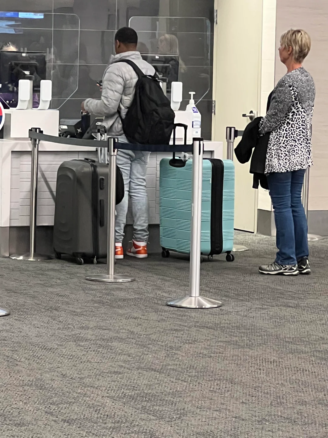 Two people with suitcases stand at an airport check-in counter. One person is wearing a gray jacket and orange shoes, the other has short blonde hair and a cheetah-print jacket. Hand sanitizer and barriers are visible on the counter.