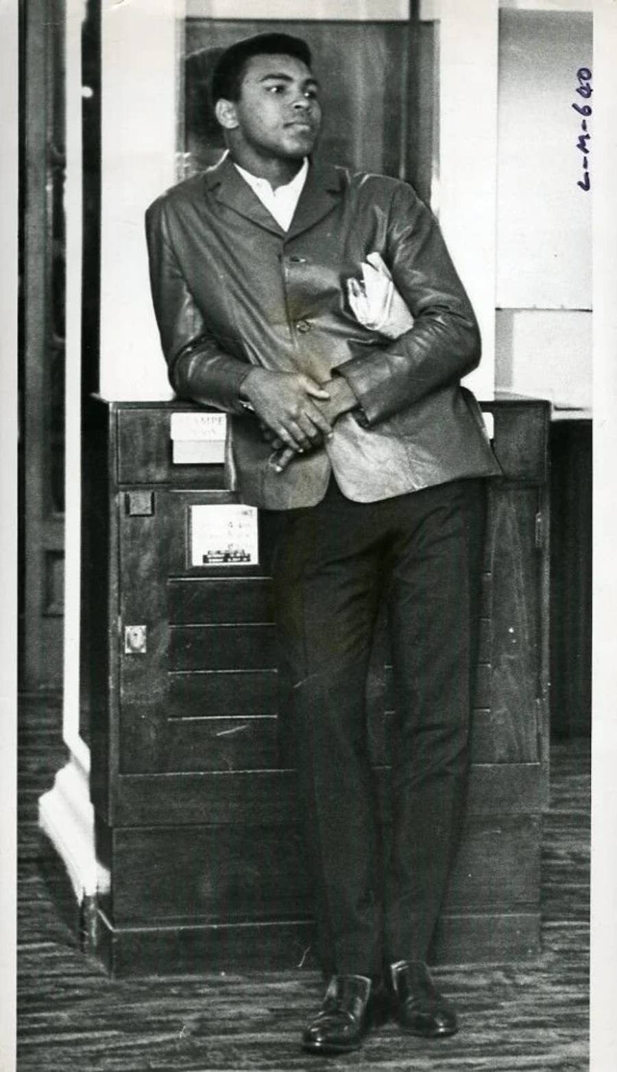 A young man in a suit leans casually against a wooden cabinet, holding papers and looking to the side in a relaxed manner.