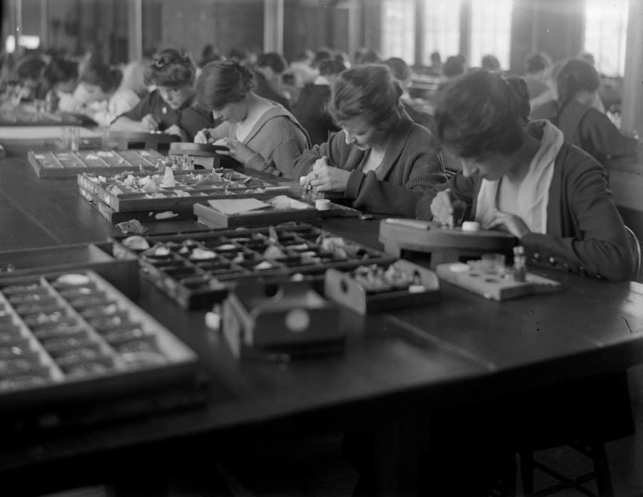 A group of women sit at long tables, closely focused on assembling or painting small objects, with trays of materials in front of them in a well-lit workshop.