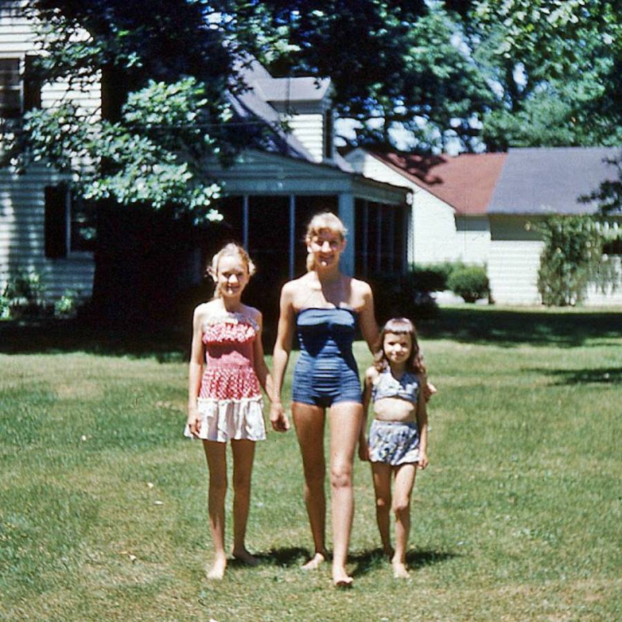 Three girls in vintage swimsuits stand barefoot on a lawn in front of a white house with green trees in the background. It appears to be a sunny day in a suburban neighborhood.