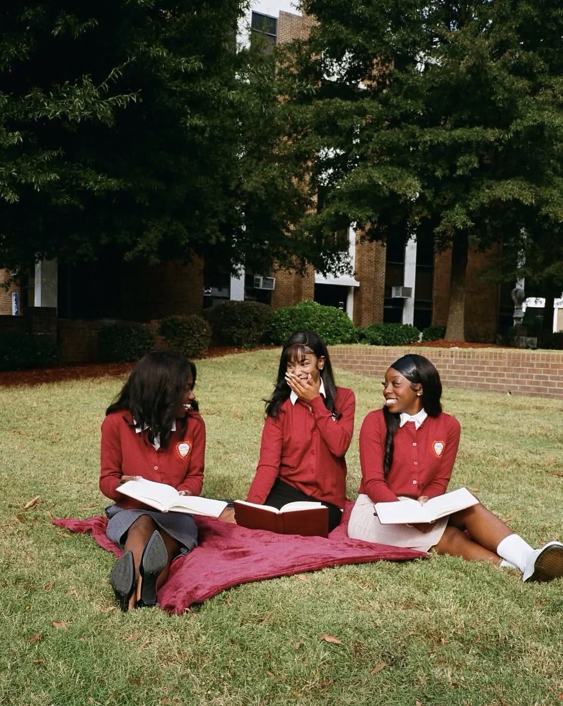 Three young women in matching red uniforms sit on a blanket outside, reading books and smiling together on a grassy area with trees and a building in the background.
