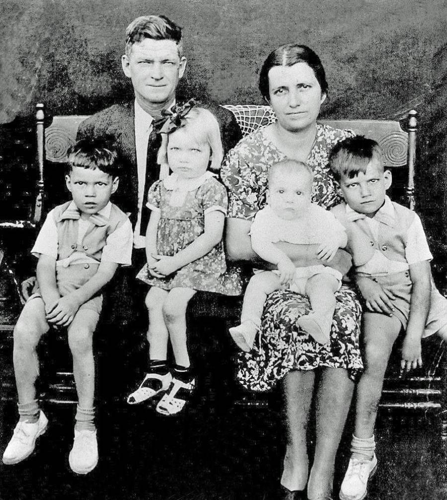 A black-and-white photo of a family of six sitting on a bench: a man and woman in the center, with three young children and a baby. Everyone is looking at the camera with neutral expressions.