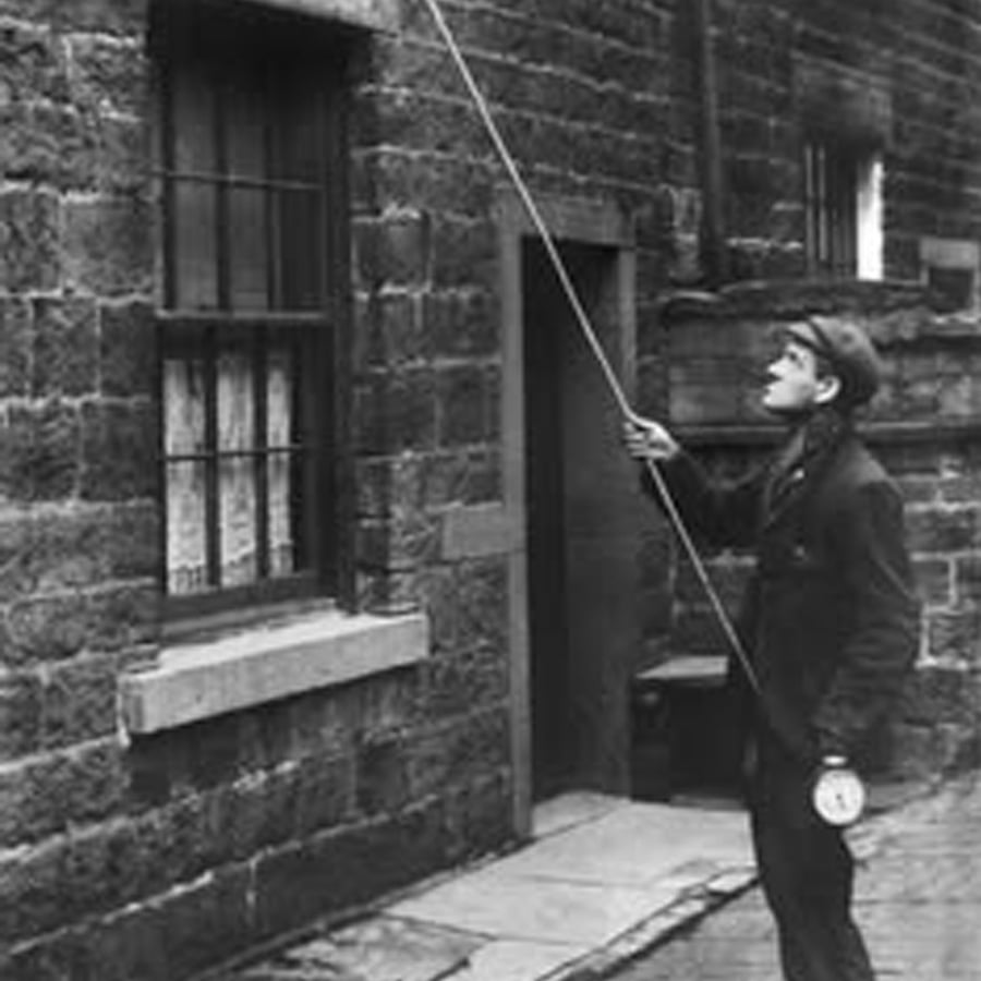 A man stands on a cobblestone street, looking up and using a long pole to tap at an upstairs window of a brick building. He wears a cap and holds a small object in his left hand.