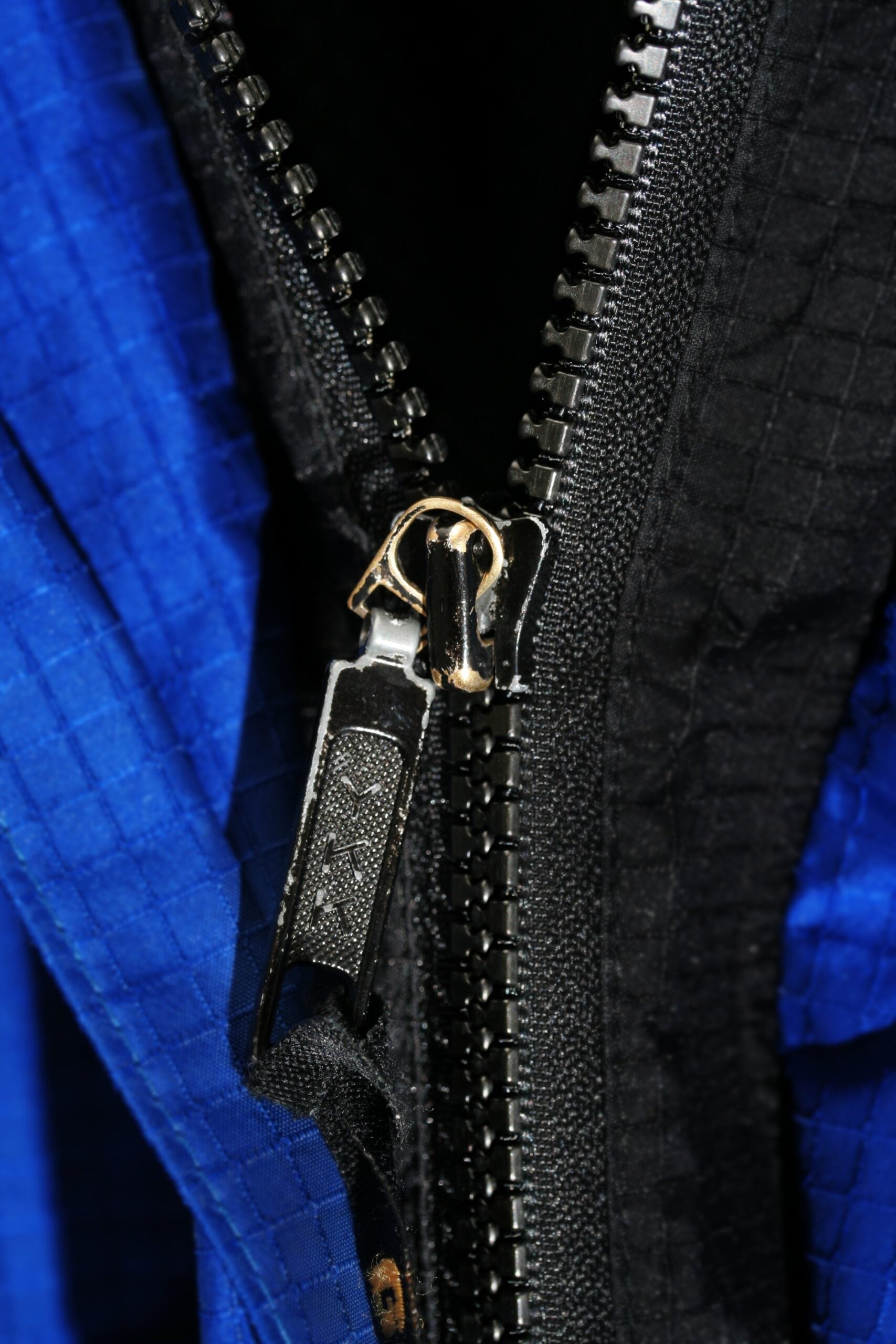 Close-up of a black zipper partially unzipped on a blue and black fabric, showing detail of the zipper teeth and pull tab against the textured material.