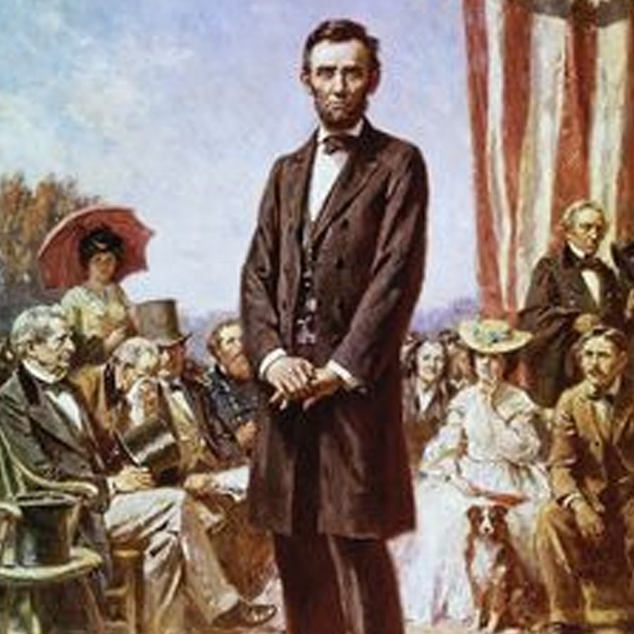 A man in a dark suit stands in front of a group of seated people outdoors, with an American flag draped in the background. Some attendees wear old-fashioned attire and hats.