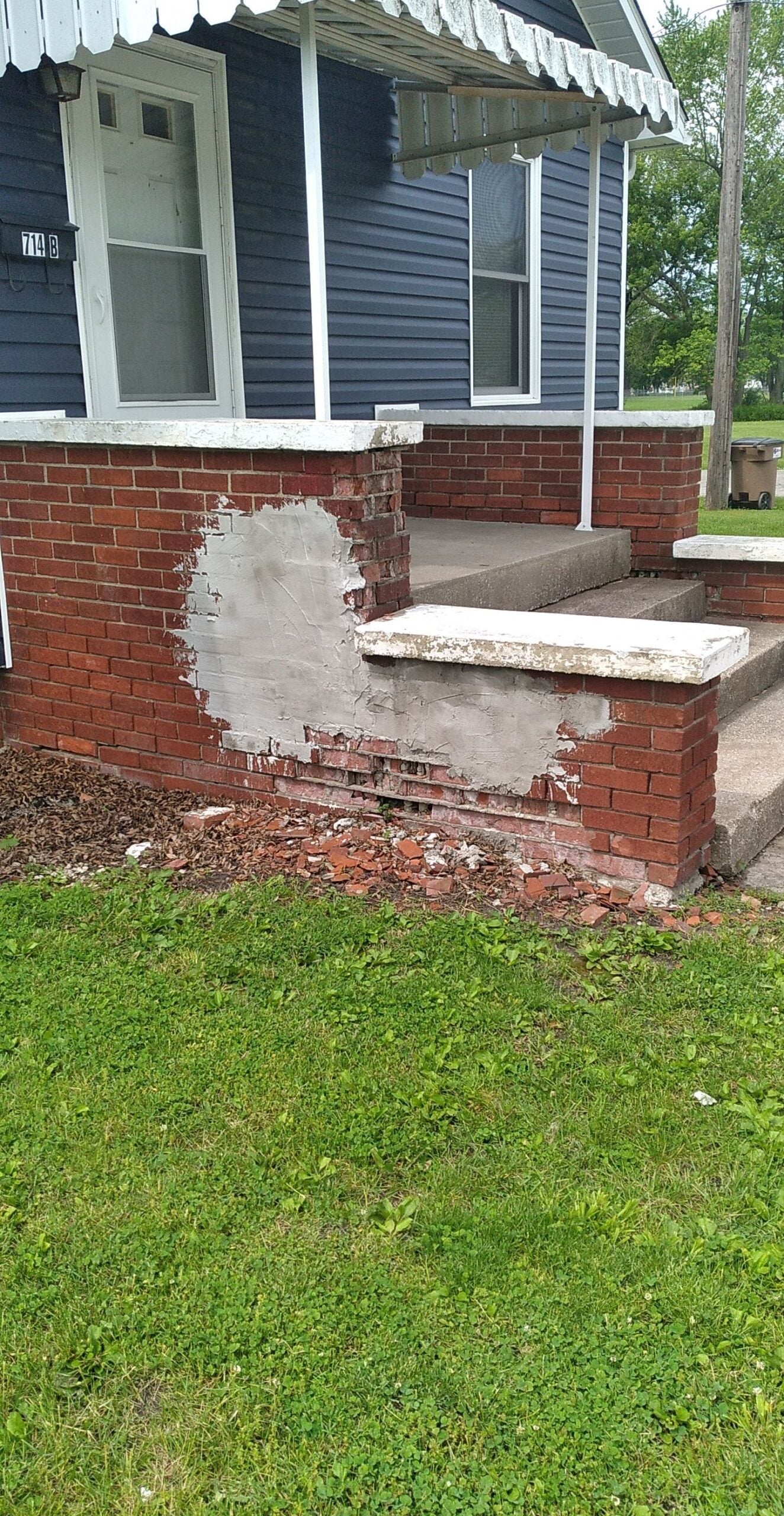 A brick porch wall with damaged and missing bricks, partially covered in gray patching cement. Some loose bricks and debris are on the ground in front of the wall.